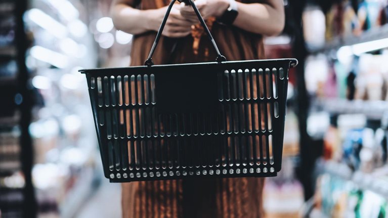 Woman standing in a grocery store aisle holding a black shopping basket.