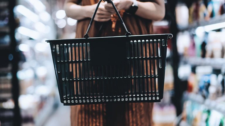 Woman standing in a grocery store aisle holding a black shopping basket. 