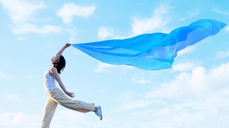A young woman joyfully leaps through the air, holding a flowing blue scarf that billows dramatically against a bright blue sky with soft clouds.