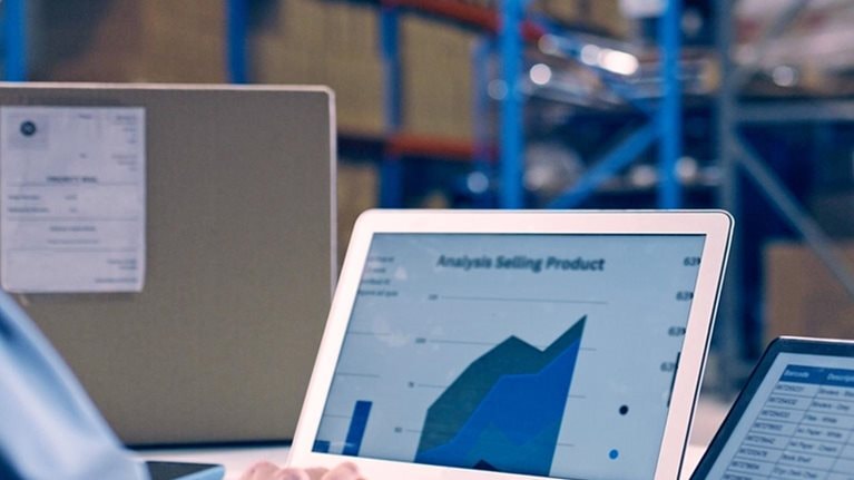 Woman working in a warehouse. Laptop screen is showing data analytics, e-commerce report and spreadsheet of sales.