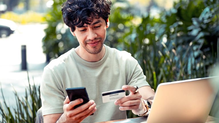 A young Latin American man sitting at an outdoor café table with a laptop. He holds a credit card in one hand and is looking at his phone in the other.