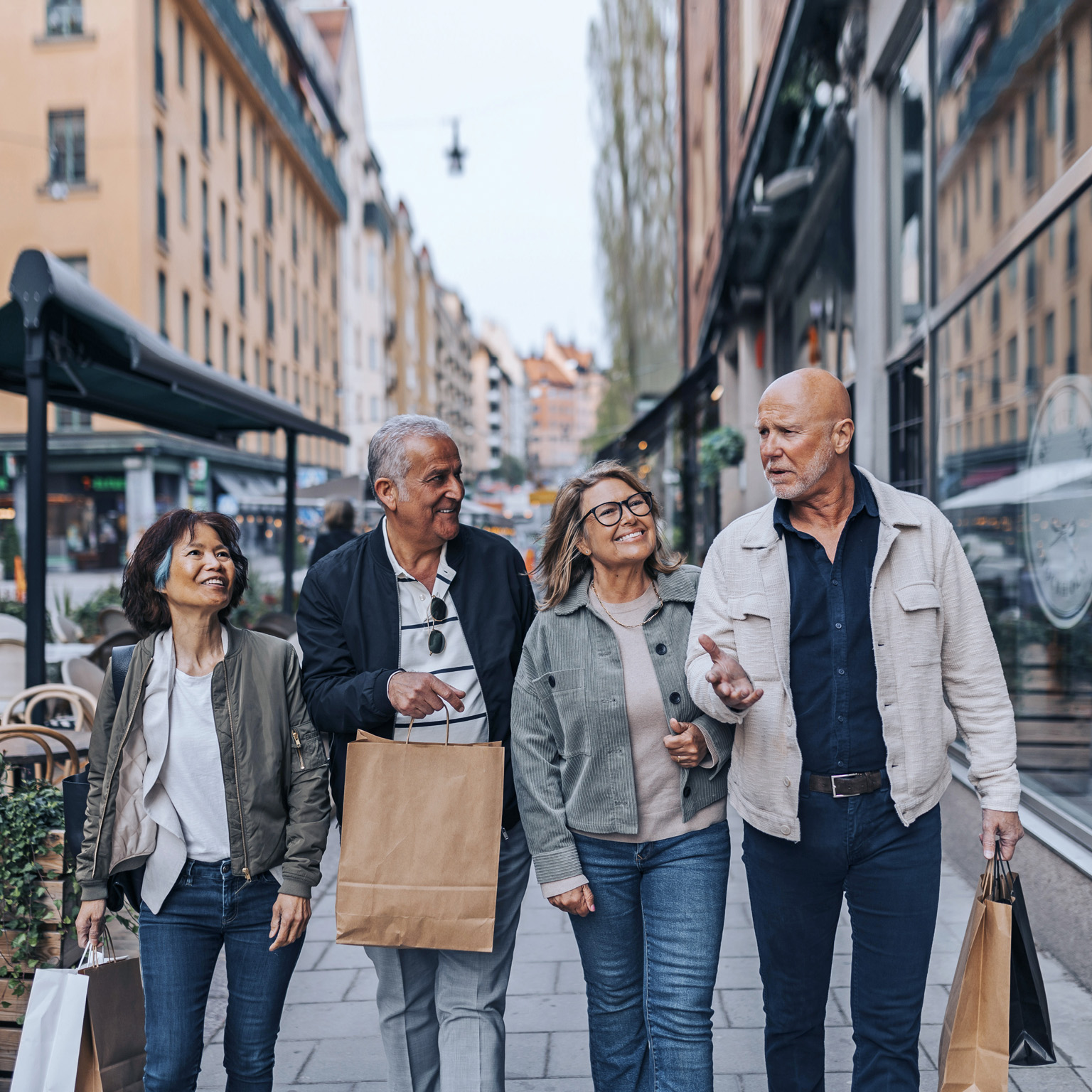A cheerful group of senior friends strolling down a European sidewalk, engaged in conversation and carrying shopping bags.