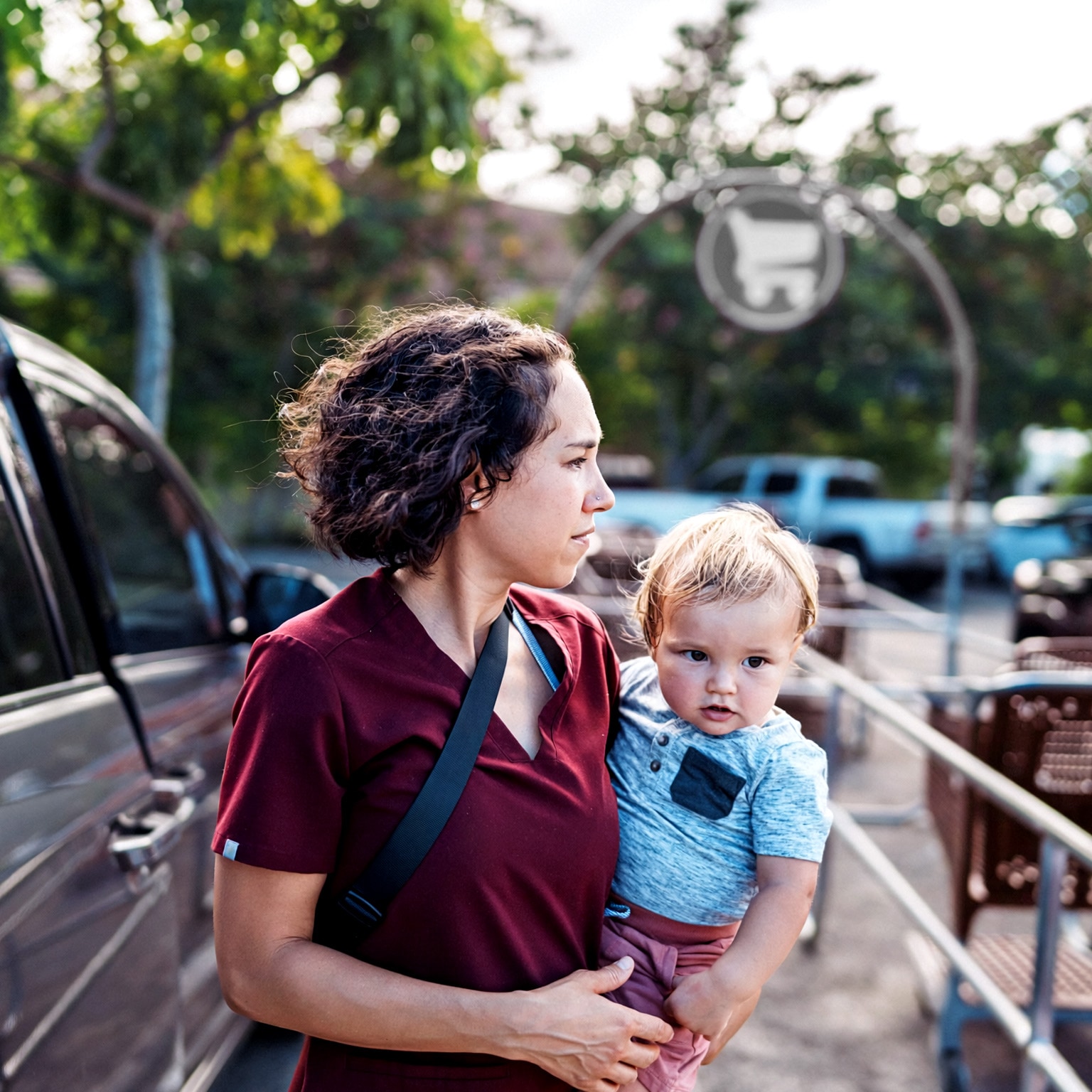 A woman holding a young child in a parking lot near shopping carts and parked cars. The woman looks off into the distance with a focused expression, while the child looks toward the camera.