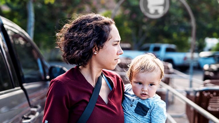 A woman holding a young child in a parking lot near shopping carts and parked cars. The woman looks off into the distance with a focused expression, while the child looks toward the camera.