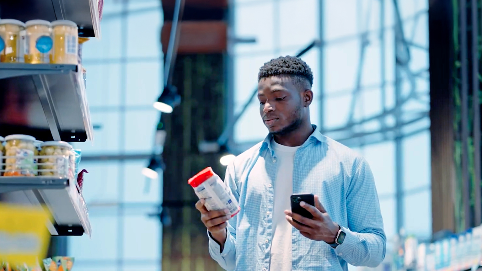 Person in a grocery store comparing a product label while holding a smartphone next to a shopping cart.