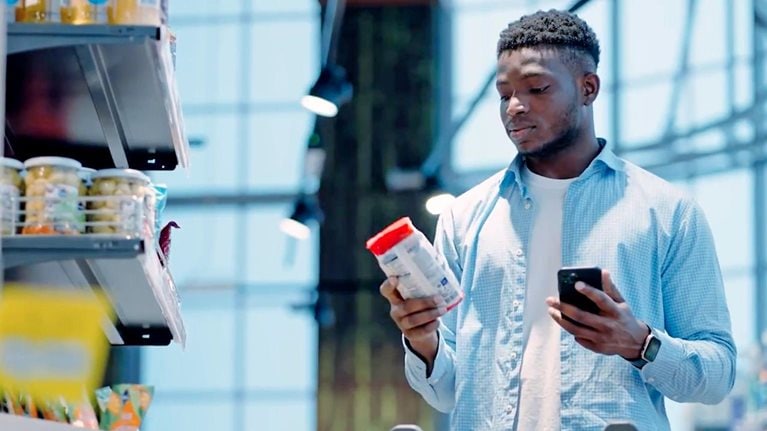 Person in a grocery store comparing a product label while holding a smartphone next to a shopping cart.