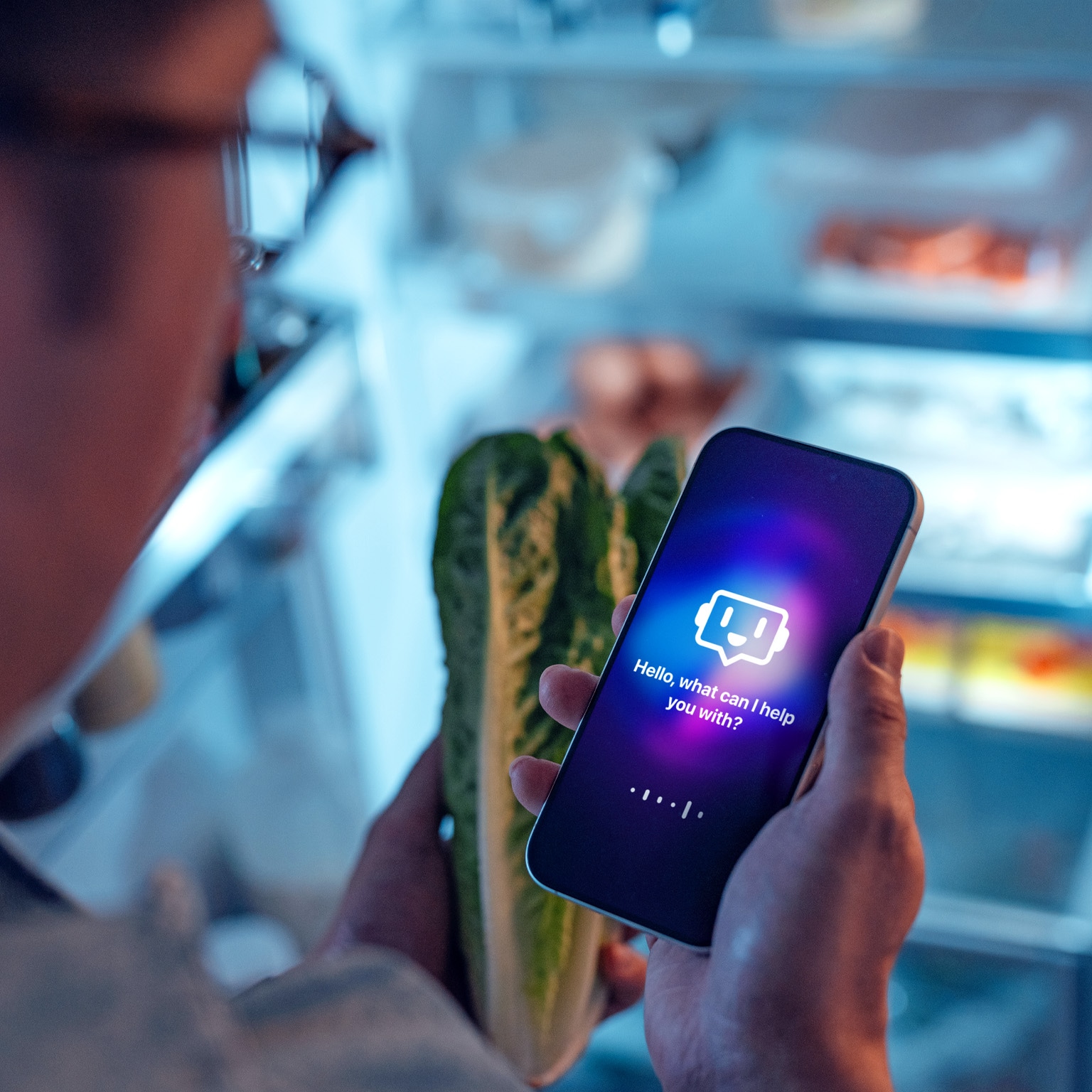 A person standing in front of an open refrigerator while holding a smartphone in one hand and a head of romaine lettuce in the other. The phone screen displays a glowing chatbot interface with the message, “Hello, what can I help you with?”.
