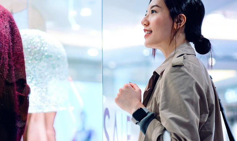 Asian woman carrying a shopping bag, standing outside a boutique looking at window display in a shopping mall