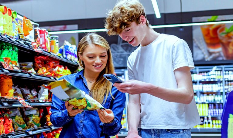 A friendly looking young couple standing in a grocery aisle with a shopping cart. The woman holds a bag of chips up to her partner while he references his phone.