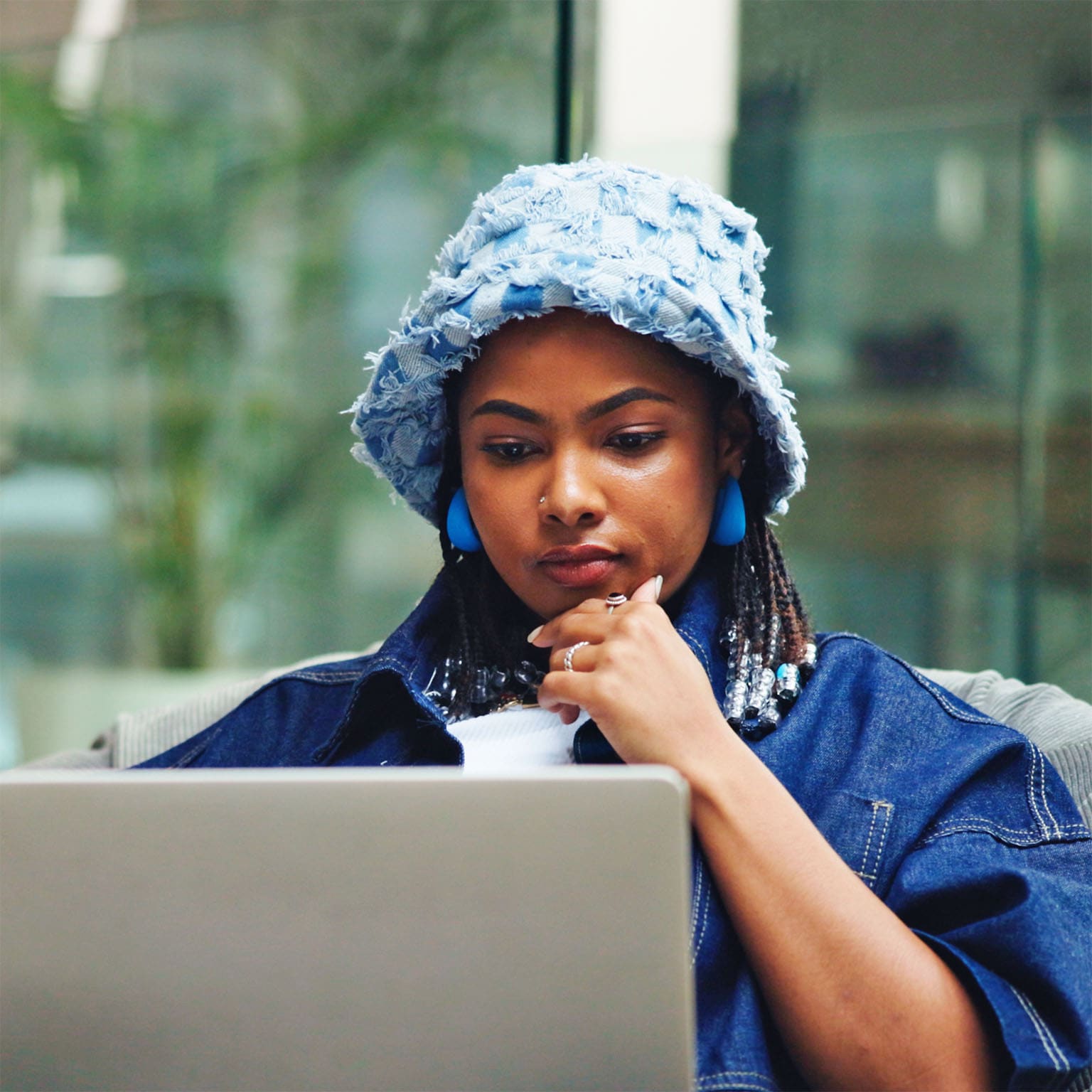 A person wearing a light blue textured bucket hat and blue earrings sits on a cushioned bean bag, concentrating on a laptop balanced on their lap with one hand resting near their chin. The background shows a modern, bright indoor space with glass walls and blurred furnishings.
