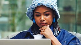 A person wearing a light blue textured bucket hat and blue earrings sits on a cushioned bean bag, concentrating on a laptop balanced on their lap with one hand resting near their chin. The background shows a modern, bright indoor space with glass walls and blurred furnishings.