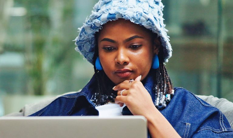 A person wearing a light blue textured bucket hat and blue earrings sits on a cushioned bean bag, concentrating on a laptop balanced on their lap with one hand resting near their chin. The background shows a modern, bright indoor space with glass walls and blurred furnishings.