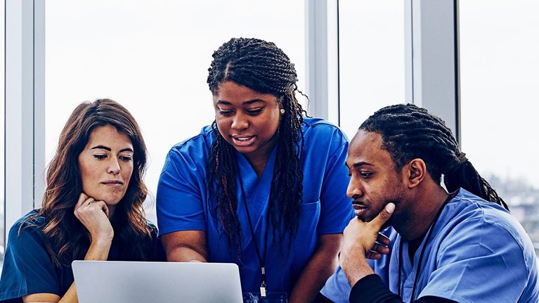 Female nurse with laptop talking to colleagues against window in hospital