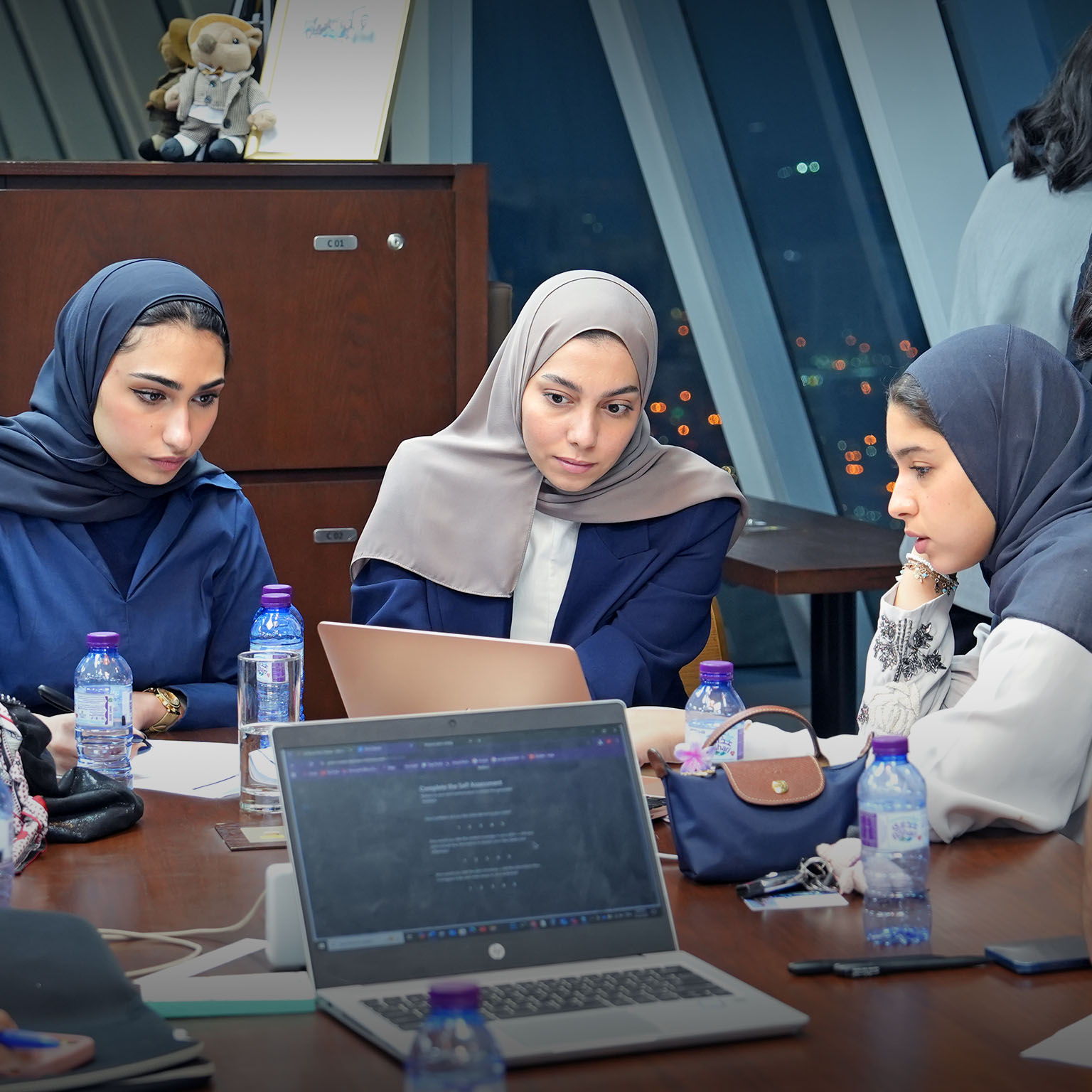 In a modern room overlooking city lights, a group of people sit around a large table with multiple laptops, notebooks, and water bottles. Several women wearing headscarves focus on a laptop while others, including a man with clasped hands, take notes or lean over to observe. 