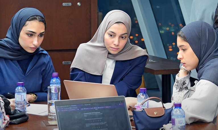 In a modern room overlooking city lights, a group of people sit around a large table with multiple laptops, notebooks, and water bottles. Several women wearing headscarves focus on a laptop while others, including a man with clasped hands, take notes or lean over to observe.