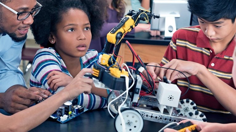 Children from diverse racial identities, who appear to be in middle school, are creating a mechanized forklift type of machine on wheels in a mechanics class with a teacher helping them. 