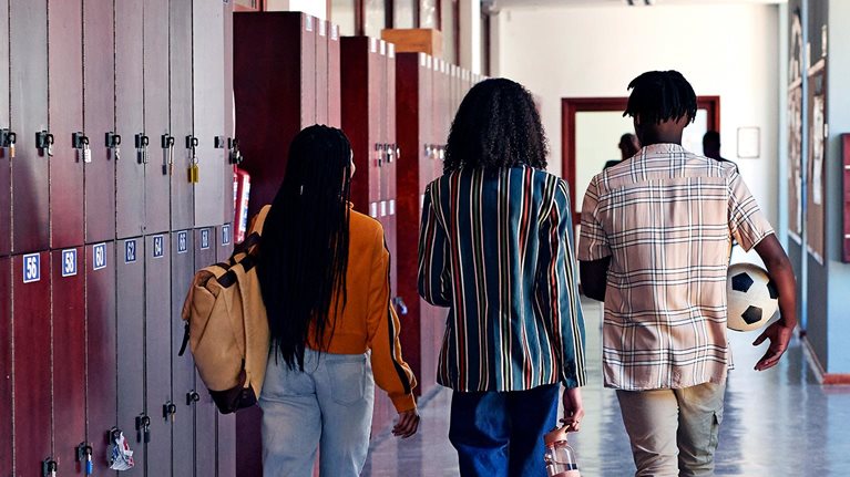 Full length rear facing view of male and female students walking together in school hallway with lockers