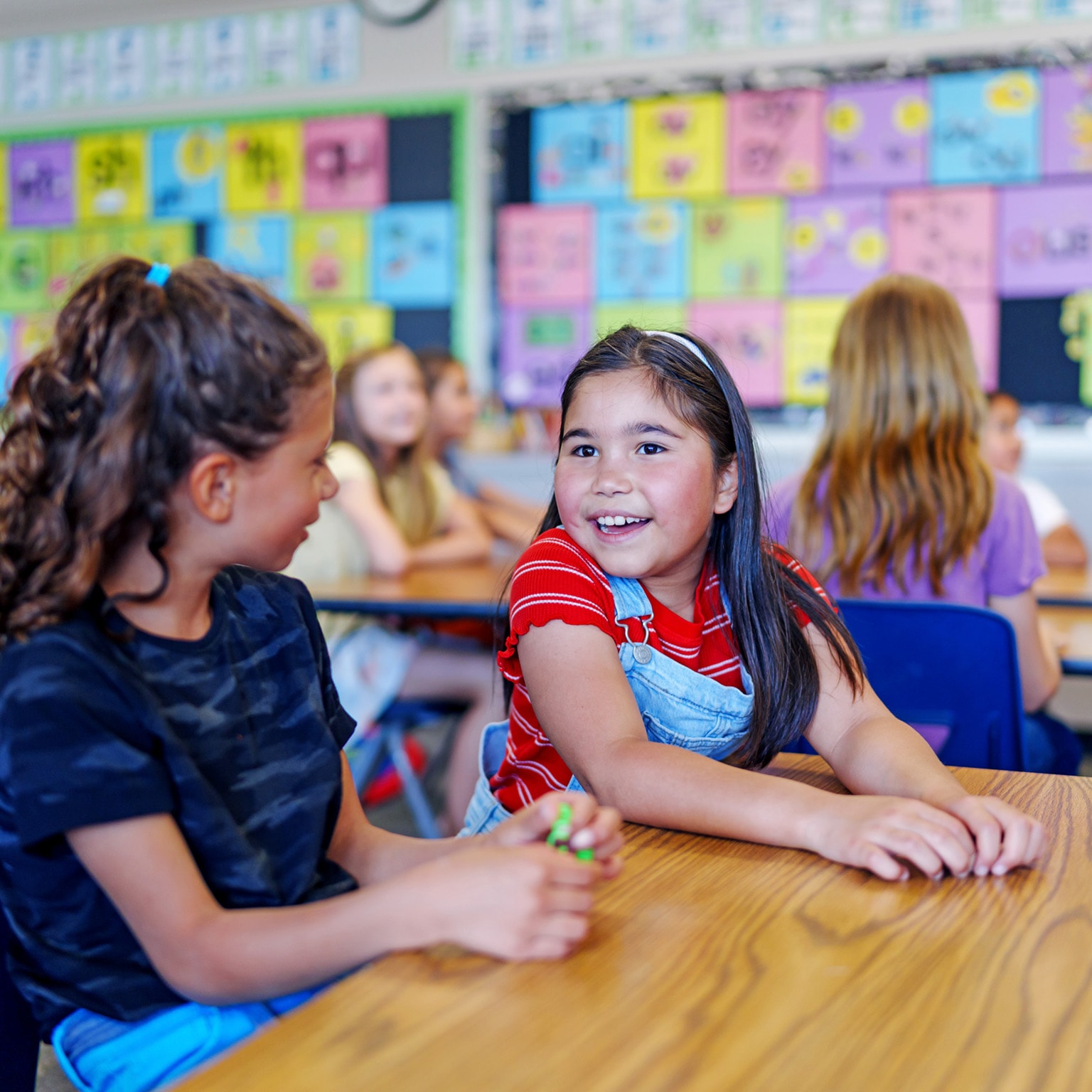 An elementary school girl sits with a friend at a desk in their classroom and talks cheerfully during class.