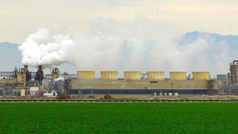 A vibrant green field in the foreground, contrasting with a large geothermal complex in the background. Smoke billows from the chimneys, and distant mountains complete the scene.