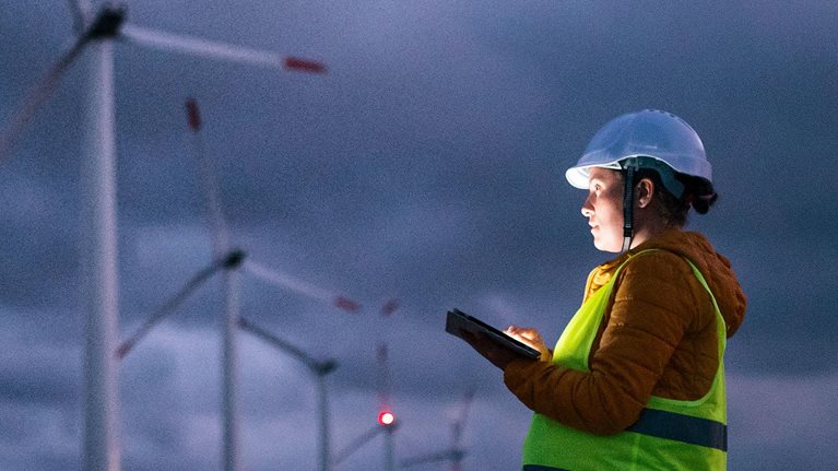 Renewable Energy Systems. Electricity Maintenance Engineer working on the field at a Wind Turbine Power station at dusk with a moody sky behind. Blurred motion. - stock photo
