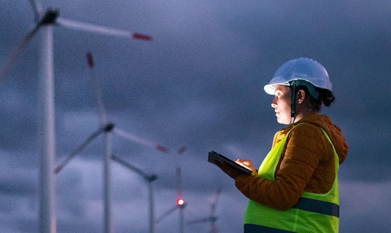 Renewable Energy Systems. Electricity Maintenance Engineer working on the field at a Wind Turbine Power station at dusk with a moody sky behind. Blurred motion. - stock photo