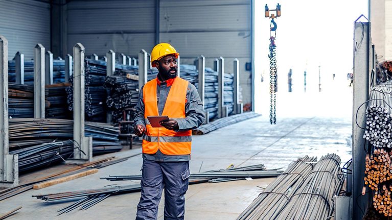 Man in a safety vest and hard hat walking through an industrial warehouse, holding a digital tablet and checking steel rebar inventory