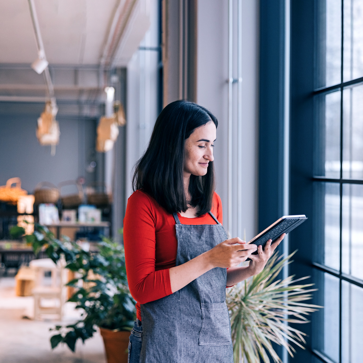 In front of a store window, a small business owner, dressed in a red shirt and apron, stands holding a tablet while glancing at it. Blurry shelves stocked with products can be seen in the background.