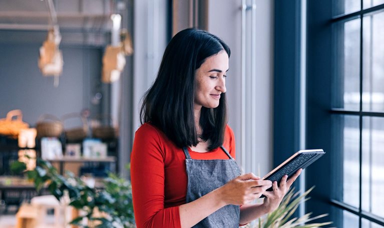 In front of a store window, a small business owner, dressed in a red shirt and apron, stands holding a tablet while glancing at it. Blurry shelves stocked with products can be seen in the background.