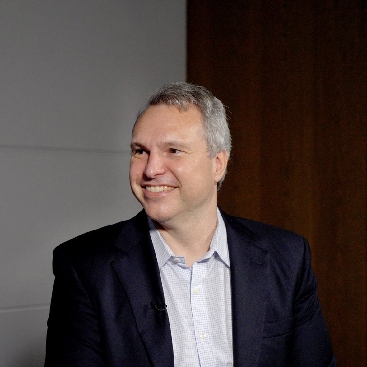 A portrait of Derek Waldron wearing a blue suit jacket over a collared shirt. He is smiling and looking to his right.
