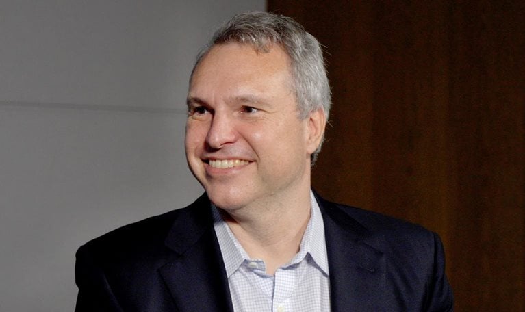 A portrait of Derek Waldron wearing a blue suit jacket over a collared shirt. He is smiling and looking to his right.