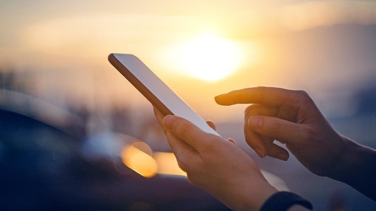 Close-up of woman's hand typing on a smartphone in the city in front of cars at beautiful sunset