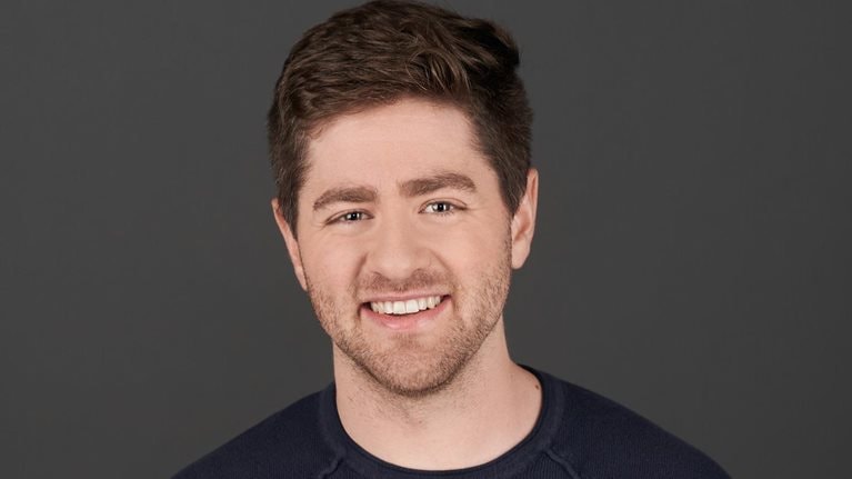 Eric Glyman with short brown hair and a trimmed beard smiling during an interview. He is wearing a dark jacket over a light-colored shirt, with a softly blurred indoor background behind him.