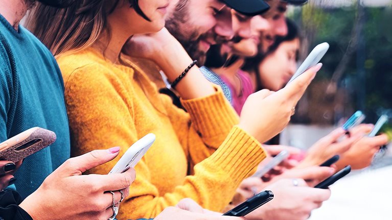 Closeup image of a group of interracial gen-z friends sitting outdoors in a row, looking at and interacting with mobile phones.