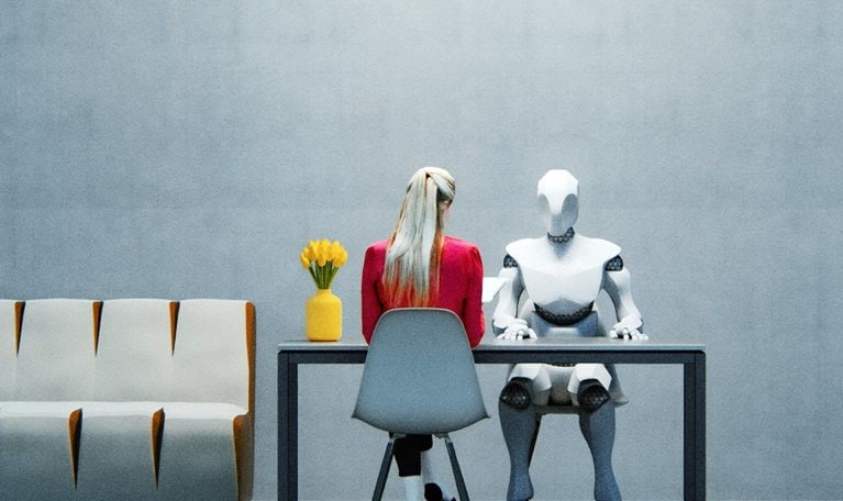 A woman in a red jacket sitting at a table across from a humanoid robot in a sparsely decorated office space.