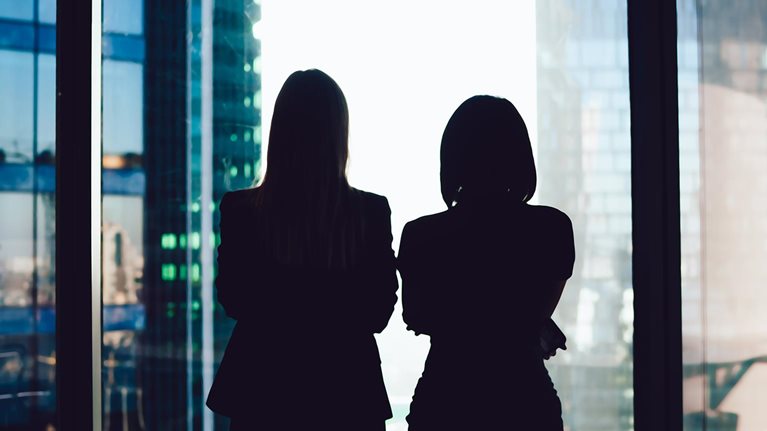 Back view of female colleagues in formal wear standing near window looking at modern exterior of skyscrapers in business center, silhouette of women together planning future success of brainstorming