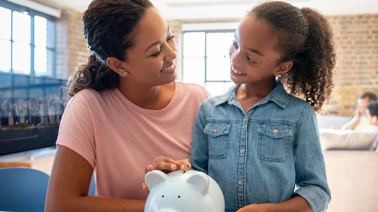 photo mother and daughter, both smiling, looking at each other as daughter drops coins into piggy bank