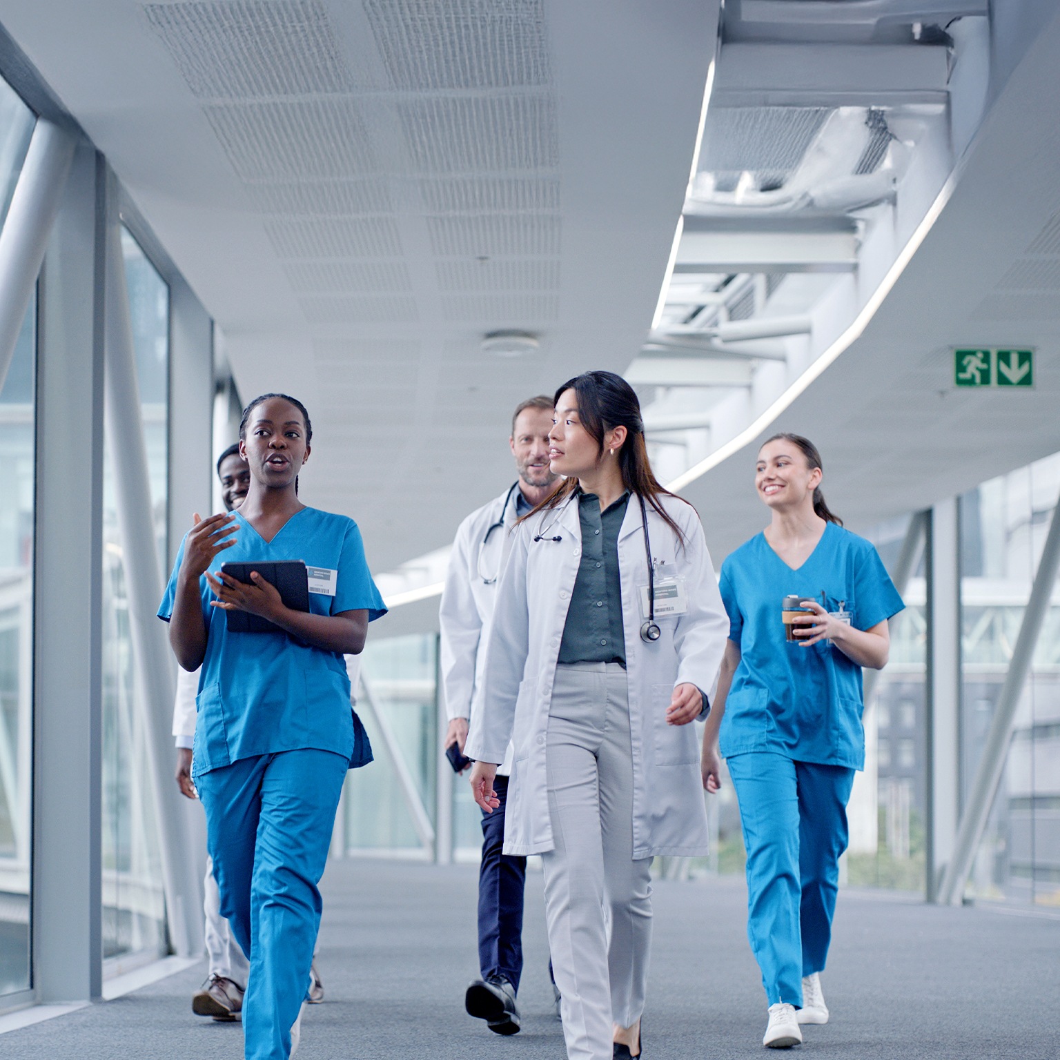 A doctor and nurse with tablet for discussion while leading a group of 5 peers that are walking down a hospital hallway together. It represents a diverse team of medical professionals utilizing technology in their collaboration. 