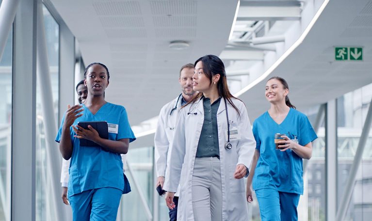 A doctor and nurse with tablet for discussion while leading a group of 5 peers that are walking down a hospital hallway together. It represents a diverse team of medical professionals utilizing technology in their collaboration.