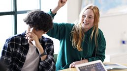 A group of students sitting at a classroom table, with a red-haired student in a green hoodie smiling and raising her hand while others, including one with a laptop, also participate.