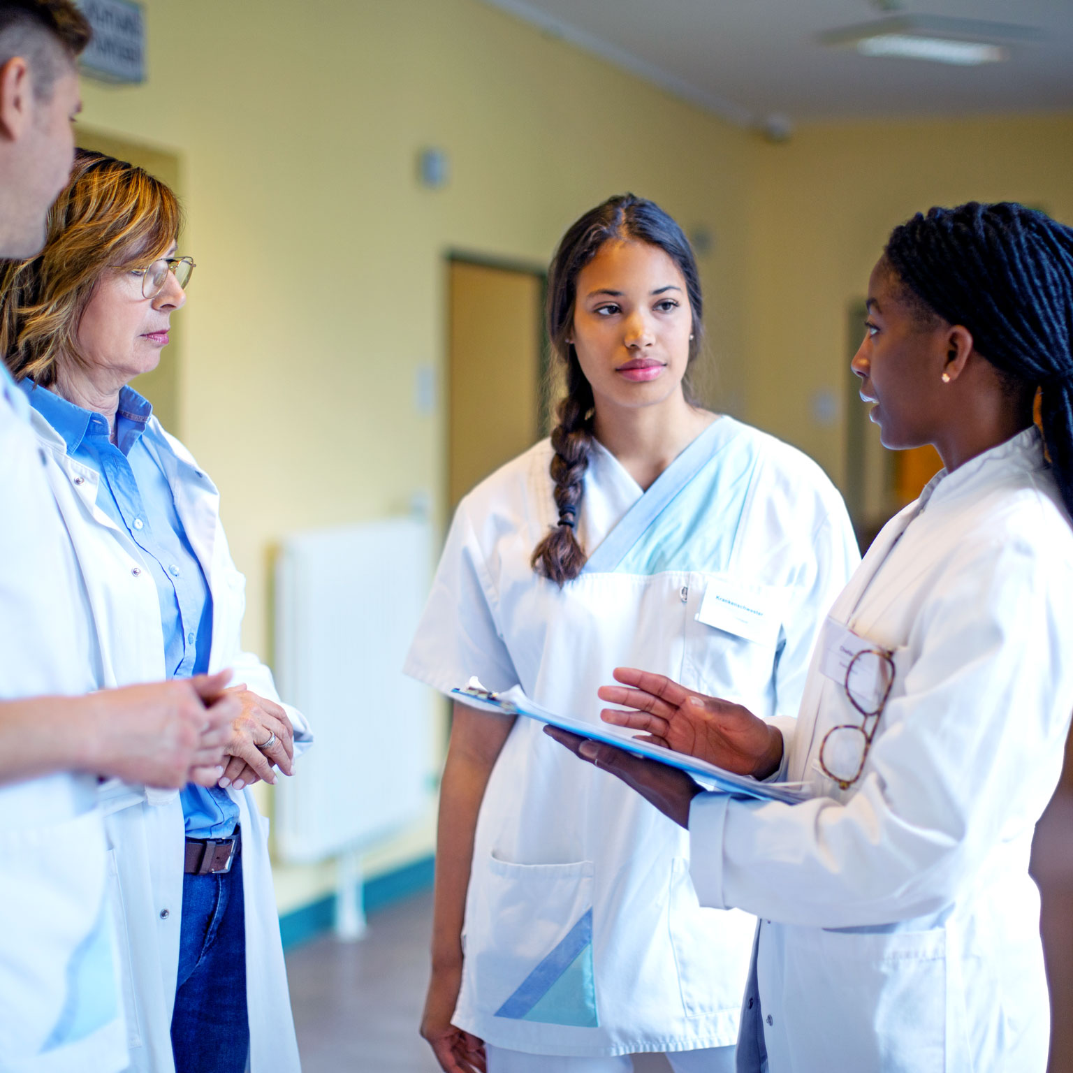 Female doctor holding medical reports while discussing with her team. Medical professionals in scrubs and lab coat meeting in hospital corridor.