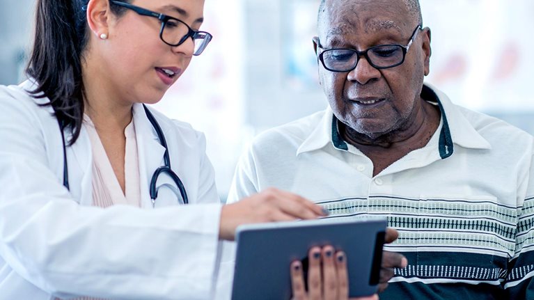 A doctor in white coat wearing a stethoscope, who is a woman, appears to explain something from an electronic tablet to an elderly black man.