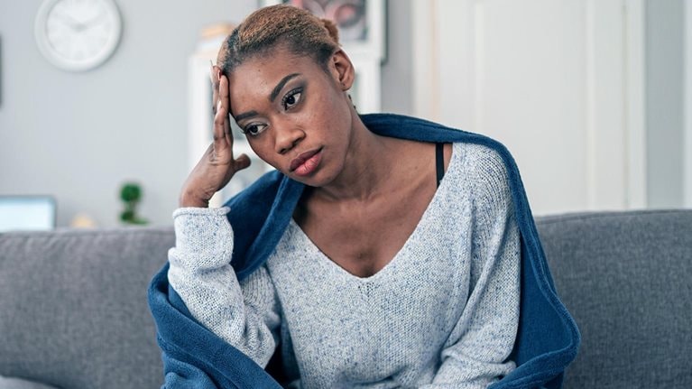 A young woman sitting on her couch wrapped in a blanket with used tissues laying next to her.