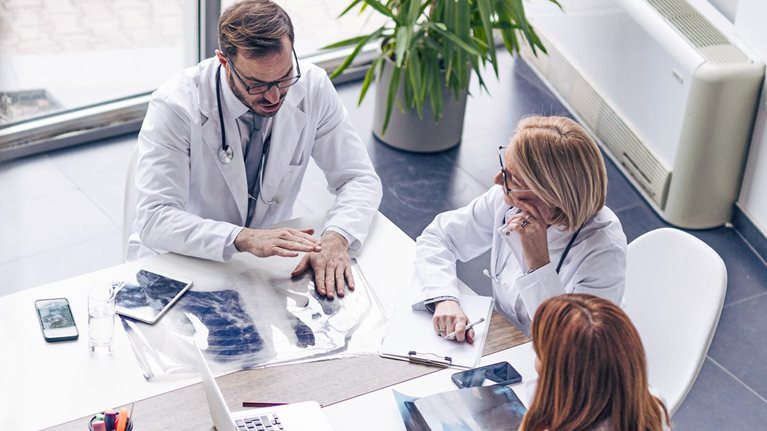 From a bird's-eye view, a diverse group of business professionals is gathered around a table covered with laptops, tablets, design plans, and financial reports. The team is engaged in a lively conversation.