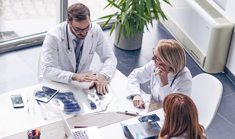 From a bird's-eye view, a diverse group of business professionals is gathered around a table covered with laptops, tablets, design plans, and financial reports. The team is engaged in a lively conversation.