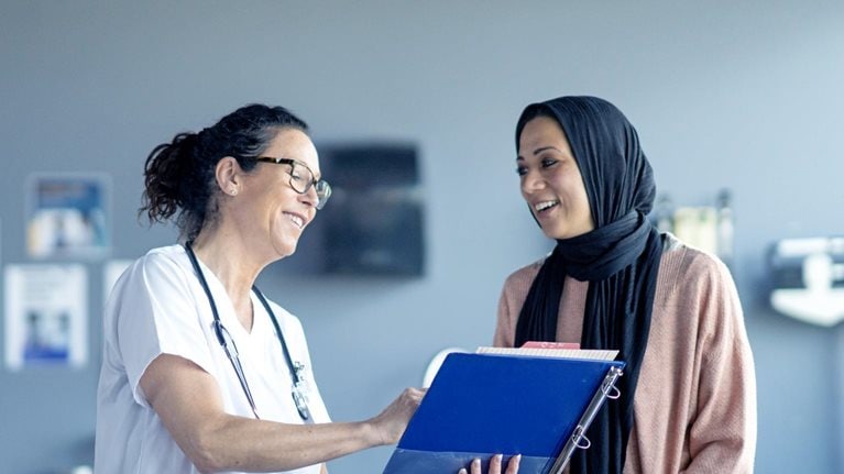 In an examination room with medical equipment behind them, a female patient wearing a black hijab and a pink sweater is greeted by a female doctor dressed in a white coat and stethoscope. The doctor holds a clipboard and smiles warmly at the patient.