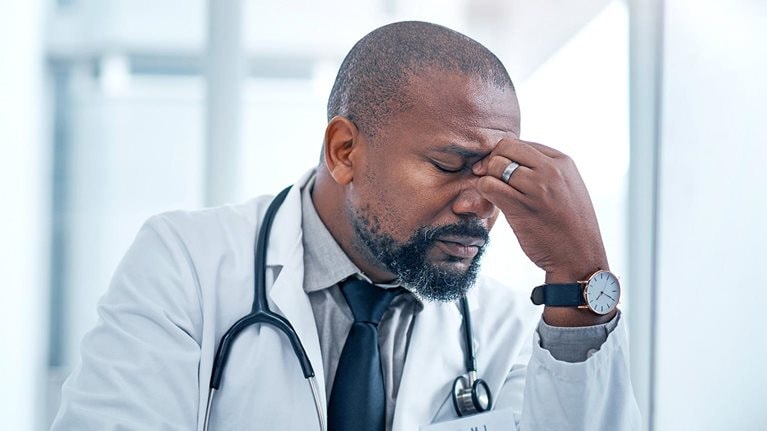 A Black male doctor sits at a computer looking frustrated