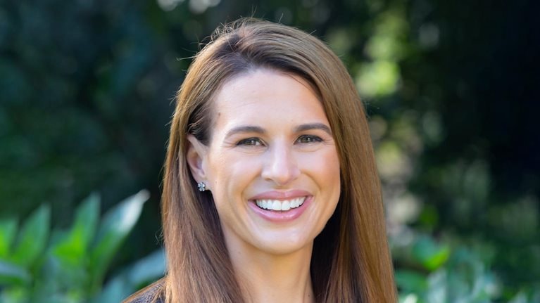 Amanda Eisel is pictured smiling, wearing a dark blue blazer against a backdrop of lush green foliage.