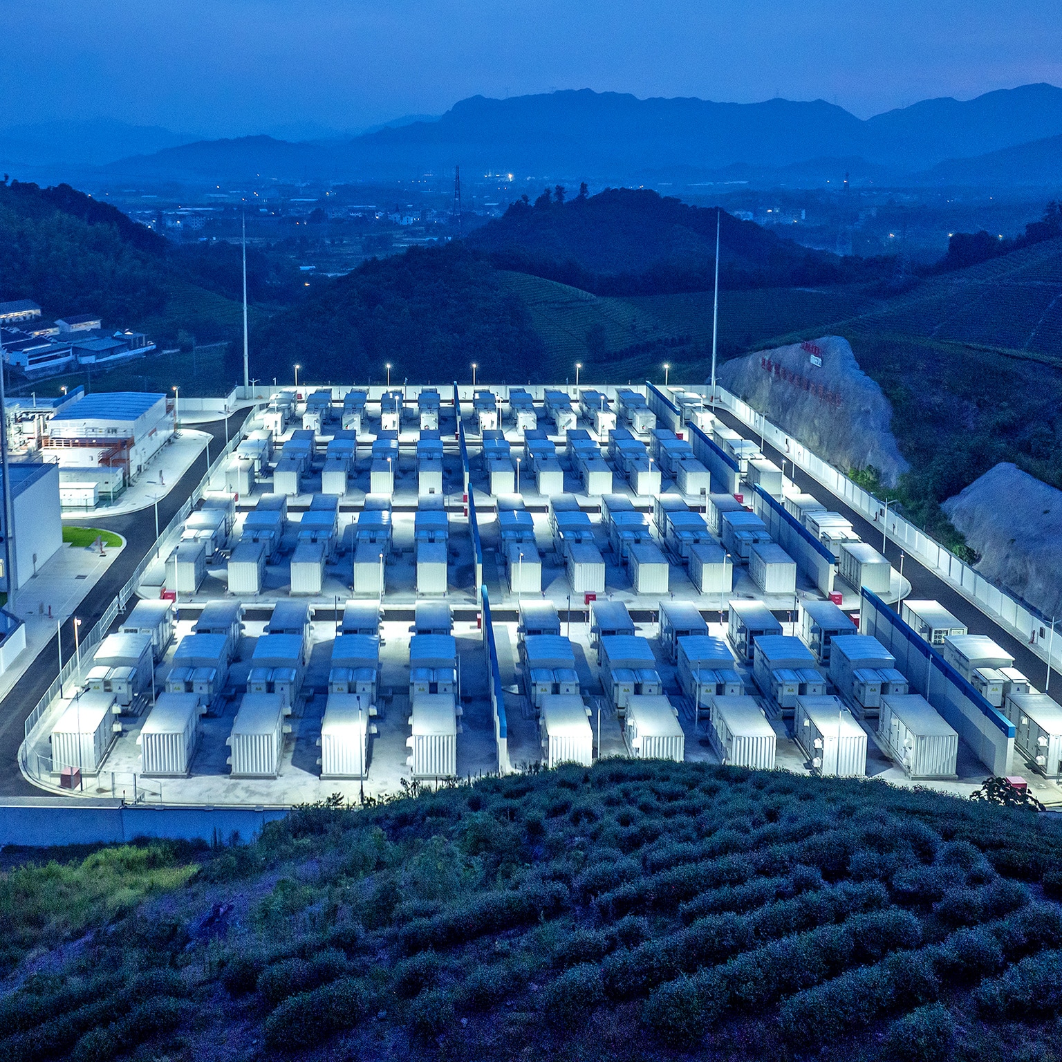 A large grid of containerized battery storage units arranged in neat rows within a brightly lit industrial facility at dusk. The site is surrounded by hills and vegetation, suggesting a large-scale energy storage installation set in a rural or semi-rural landscape.