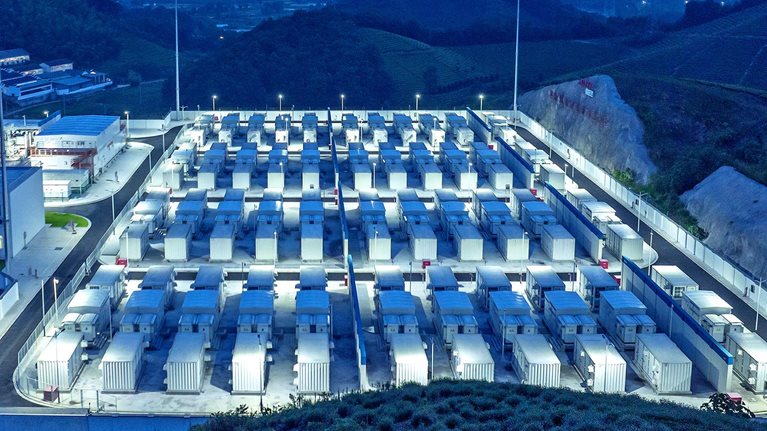 A large grid of containerized battery storage units arranged in neat rows within a brightly lit industrial facility at dusk. The site is surrounded by hills and vegetation, suggesting a large-scale energy storage installation set in a rural or semi-rural landscape.