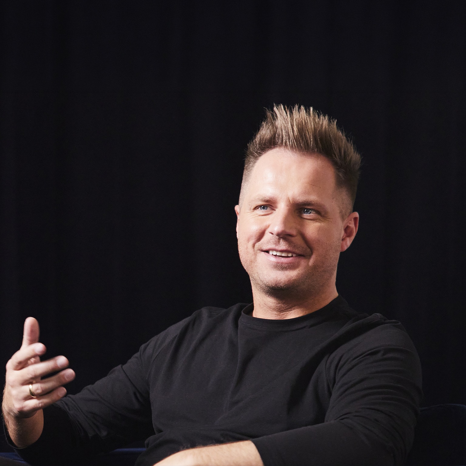 Photo of David Reger sitting on a couch on a stage, and gesturing with his hand while sharing an idea.